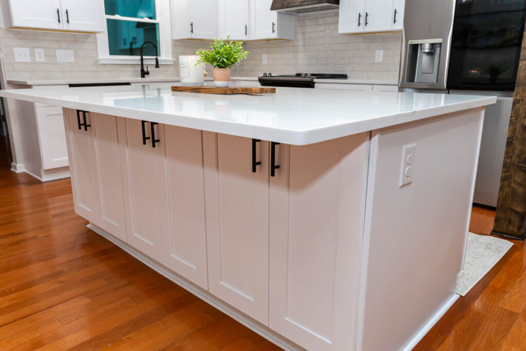 White kitchen island with black handles, white countertop, and small decor items on top, set on a wooden floor.