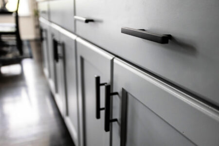 Close-up of gray kitchen cabinets with black metal handles and drawers, set against a dark reflective floor.